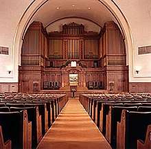 Facing the viewer are the backs of many rows of pews, with a carpeted aisle in the center. At the front of the room is a very large, wooden structure, which fills the far wall.