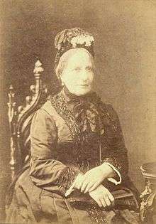 A photographic portrait of a seated woman with graying hair who is dressed in a dark and elaborate late-Victorian style dress and wearing a flowered bonnet