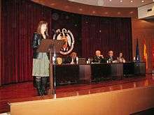 Woman speaking onstage, with others seated at a dais