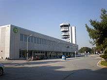 A large, square, two-story, gray building with "T2" on a sign, in front of a street. In the background is a control tower