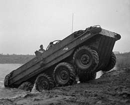 A large, open-topped, eight-wheeled vehicle emerges from a body of water and is climbing up the bank and onto land; two men are on board it.