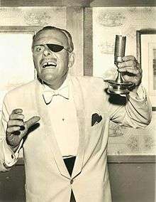 Terry-Thomas wearing a white dinner jacket. He wears an eye-patch and carries a candle