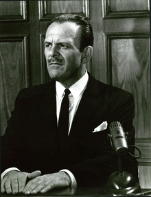 An unsmiling Terry-Thomas wearing a dark suit and tie in a witness box