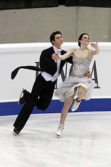 A man and a woman ice dancing; the man, on the left, is wearing black tie and tails; the woman is wearing an off-white dress with a fluffy skirt.