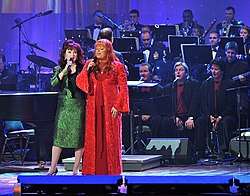 Two women, one dressed in red and one in green, singing on stage in front of an orchestra