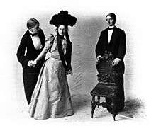 Three female actors, with two dressed as men and the third, in the center, dressed in an elegant gown with a large headpiece; the leftmost character offers a chair, being held by the rightmost character, to the woman in the center.