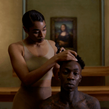 A black woman with short hair giving a shirtless tattooed black man a haircut in the Louvre art museum. Leonardo da Vinci's Mona Lisa can be seen hanging in the background.