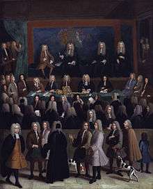 A rectangle picture of a courtroom.  Dozens of men in a courtroom in 1750s era court suits and wigs.  A blue wall at the back contains a coat of arms.  On a raised stage at the back are four men.  Several onlookers, some with dogs and children, pass by on a sidewalk looking in on the court proceedings.