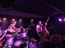 Three men are seen playing. The man at left is almost completely cut off and is playing a guitar. The second man is shown in right profile, he stands behind the drum kit and has a microphone near his face; he holds a single drum stick and is shirtless with blurred tattoos on his upper right arm. The third man is also shirtless with a microphone near his face; he is playing an upright bass. A fourth man is difficult to see in the dark, he appears to wear a skull mask.