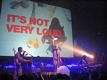 A rock band performs on stage with a graphic that says "IT'S NOT VERY LOUD" is on a screen behind them.