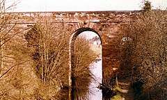 A tall, brown stone bridge with one wide central arch over a canal, and a secondary arch over a towpath. The bridge is partially obscured by trees.