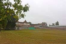 A grass playing field with buildings and tennis courts in the background