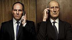 Head and shoulders of two serious-looking men in dark suits standing side-by-side, facing the camera, in front of dark wooden panels, each holding a phone to their ear.