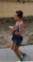 A woman with short brown hair in running motion, wearing a gray tank top and darker gray tight-fitting shorts and neon blue and green running shoes. She is on a sidewalk with a cream-colored wooden fence behind her