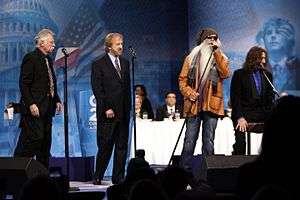 Four men standing at microphones on a stage.  Three are wearing black suits.  The fourth is wearing a tan jacket, dark glasses and jeans and has an extremely long white beard.