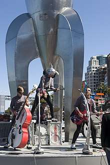 Four band members in grey and black uniforms playing in front of a metal rocket sculpture.