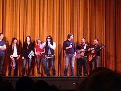 Wiseau and Sestero with microphones on the theatre stage with multiple musicians behind them.