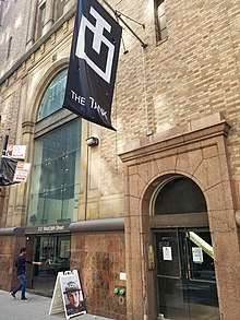 A yellow brick building with two double glass doors, one to the left, the other to the right, runs out of the frame at the top. A person is entering through the left glass doors. A black and white banner hangs from the second story with The Tank's logo and the words "THE TANK".