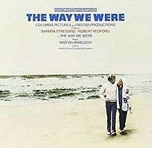 Actors Barbra Streisand and Robert Redford walk on a beach barefoot.