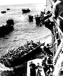 Marines board landing craft from a transport ship while another landing craft moves away from the ship