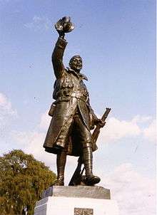 Statue of a soldier with rifle at his left side and hat raised aloft in his right hand.