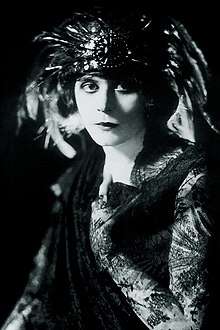 Black and white photo of a white woman wearing a feathered headdress and a dark gown with lace sleeves