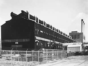 A large dark-coloured rectangular building and a smaller building with three smokestacks