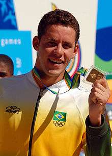 Brazilian athlete poses for photographs after the awards ceremony of the men's 200 metre individual medley competition. Wearing the official uniform of the Brazilian delegation, he holds the gold medal with his left hand.