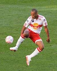 A coloured photograph of a footballer, wearing a white shirt and red kit, adorned with white socks and boots. He is controlling the ball with his right leg from an elevated position.