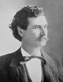 A man with bushy, black hair and a thin mustache wearing a black jacket, vest, and tie and a white shirt