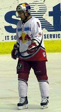 An ice hockey player standing, looking to the left of the camera. He is wearing a blue helmet and a red and white uniform.