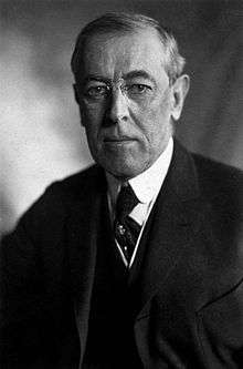 Half-length portrait of a man wearing a three-piece suit, a necktie, round-rimmed glasses and looking directly towards the photographer with an emotionless expression.