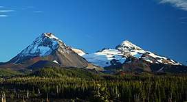Two volcanoes rise above a landscape of mixed forest and lava, with a glacier spanning the area between the two peaks
