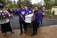 A group of six people dressed in purple carry a white coffin from concrete onto a footpath, next to a woman in a white shirt holding a bouquet of flowers.