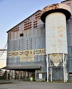 Photo of a rusted structure built of corrugated sheet metal, painted with faded letters reading "Tichnor Dryer & Storage", sitting just behind a tall, cylindrical rice storage elevator.