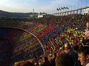 A crowd of spectators in an open-air stadium, a mosiac in red, blue and gold is visible