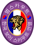 An oval badge of red with 범스카우트 (Tiger Scout) and 한국 보이 스카우트 연맹 inscribed in hangul; The center is an oval of yellow with the profile of a tiger head over a scroll inscribed with "preparation" and knot