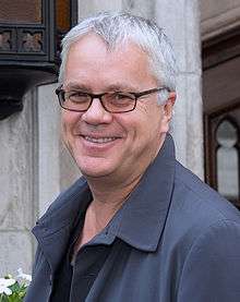 A standing caucasian man with short white and grey hair, wears glasses and a blue coat. He faces left towards the camera smiling.