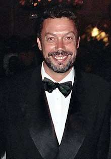 Tim Curry, a Caucasian man with a beard and short brown hair facing the viewer and smiling. He is wearing a Tuxedo.