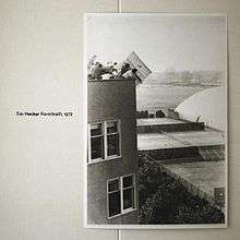 A white wall with a black-and-white photo of students dropping an upright piano off the roof of MIT's Baker House. The text on the left side reads "Tim Hecker Ravedeath, 1972".