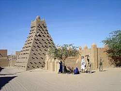A street with a mud wall and a pyramid shaped mud building with sticks protruding from its wall.