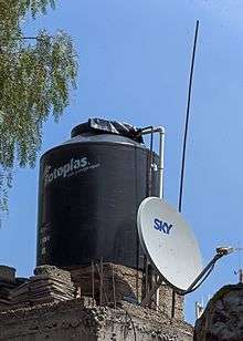 A black cylindrical tank and gray satellite television dish on a concrete roof in front of blue sky with a willow branch at top left