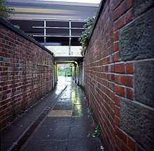 Narrow lane between two high brick walls leading to a tunnel under a railway bridge.