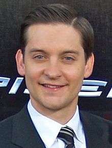 An image of a Caucasian man smiling. He has medium brown hair and is wearing a navy jacket over a white shirt and striped tie.