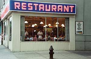 The exterior of a restaurant at the corner of a street. Through the windows a waiter can be seen taking orders. Above the windows is the word "Restaurant" in big pink letters.