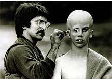 Savini, on the left, applies make-up behind Lehman's ear. Lehman's bald head has been made to appear over-large; his eyes point in  directions, and his teeth are extremely crooked.