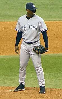 A dark-skinned man in a gray baseball uniform with "New York" across the chest in navy-blue letters and a navy-blue baseball cap standing on a dirt mound on a grass field and wearing a black baseball glove on his left hand