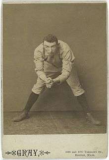 A black-and-white photograph of a man in a white baseball uniform bent over from the waist but looking into the camera