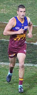 A man in a maroon, blue and gold football jersey and maroon shorts jogs on grass