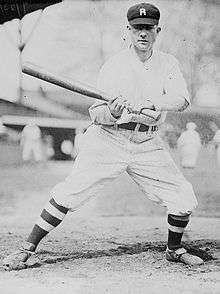 A man in a white baseball uniform with dark pinstripes, high dark socks, and a dark cap with a "R" on the front stands leaning to the right with his bat out to his left side.